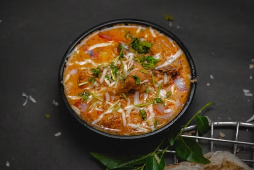 A bowl of creamy Indian curry garnished with chopped onions, coriander leaves, grated coconut, and curry leaves, served in a black bowl against a dark background